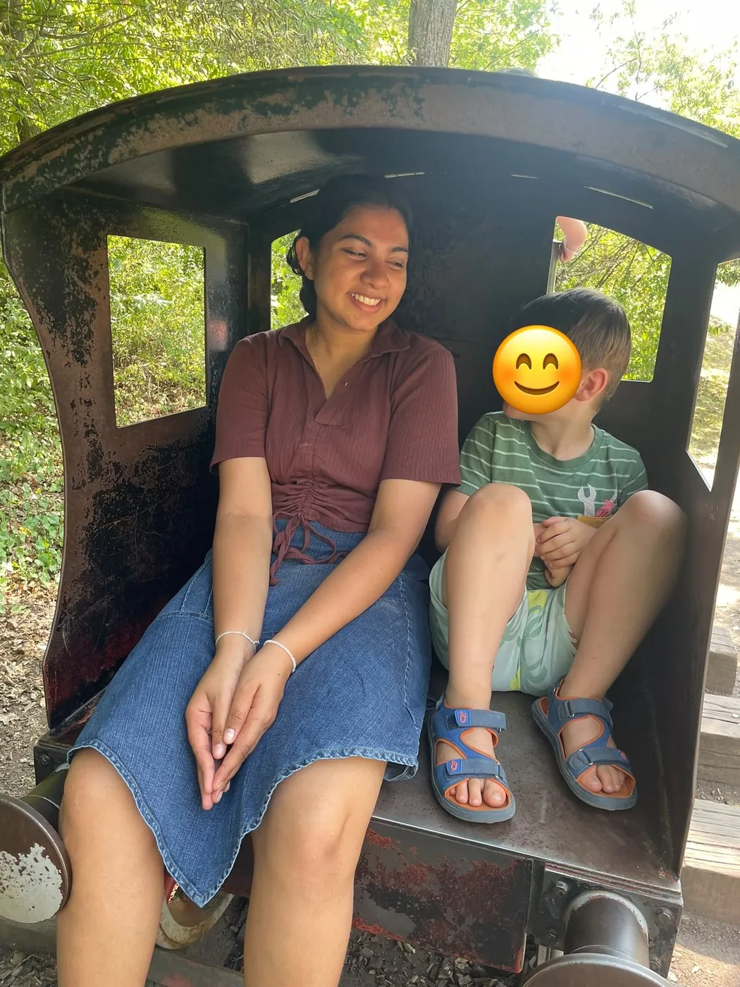 Au-pair und Kind sitzen im Sommer nebeneinander in einem Waggon einer Parkeisenbahn; das Gesicht des Kindes ist mit einem Smiley abgedeckt.